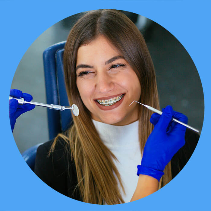 A smiling woman with braces on her teeth during a dental check-up, with a dentist's hands in blue gloves holding tools near her mouth.
