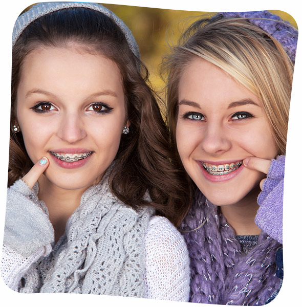 Two smiling teenage girls wearing winter clothes and pointing to their braces.